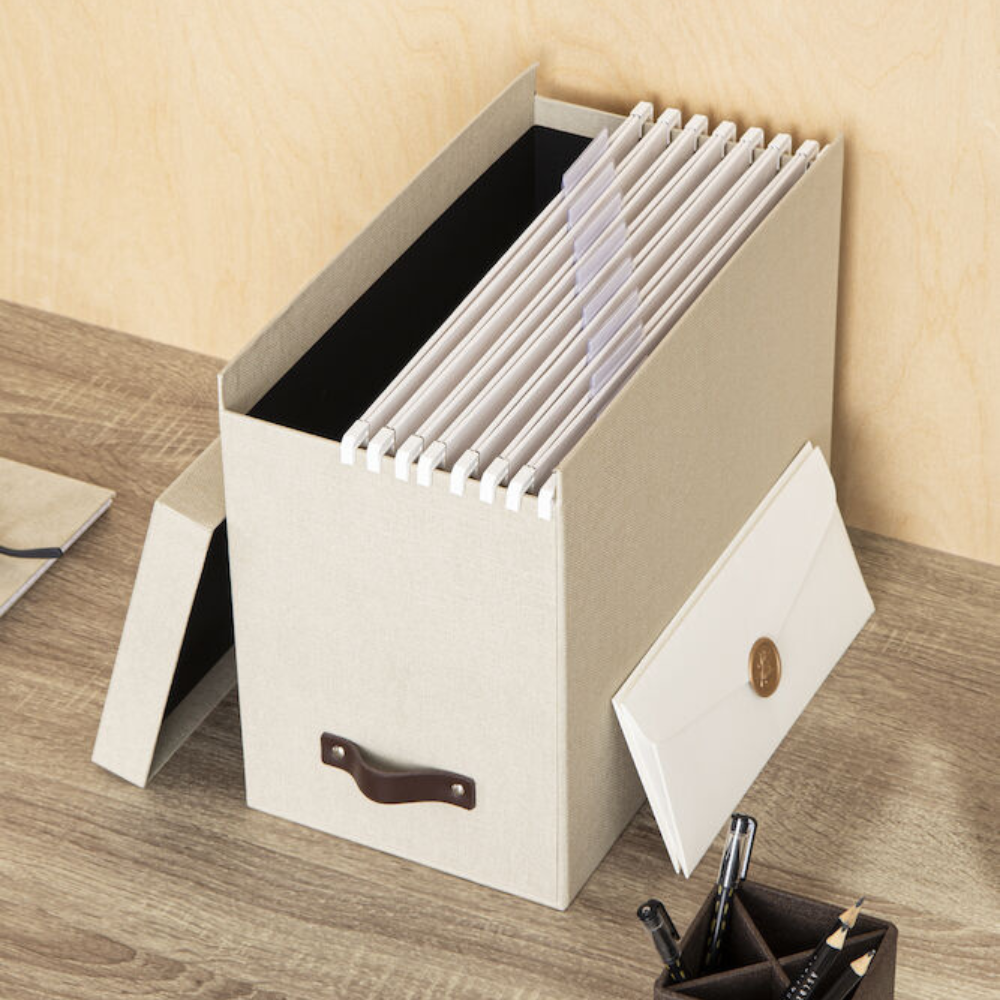 De Bigso JOHAN hangmappenbox van linnen canvas met een bruine handgreep staat op een houten bureau en biedt plaats aan A4-mappen en een envelop. Er liggen schrijfwaren bij voor het eenvoudig ordenen van documenten.