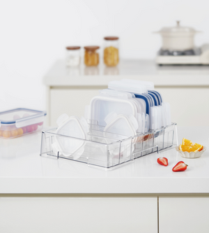 Op een aanrecht staat de BNB x VCN Dekselorganizer KLAR – met flexibele vakken waarin deksels en plastic bakjes netjes worden opgeborgen. Ernaast liggen stukjes fruit, een bakje met deksel, en op de achtergrond staan potjes en een pan.