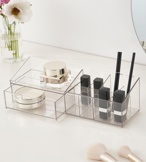 De doorzichtige multi-organizer KLAR (30,6 x 9 x 9 cm) van BNB x VCN houdt de gouden potjes, zwarte lippenstiften en make-upborstels netjes op hun plek op de witte kaptafel, naast de vaas en de ronde spiegel.