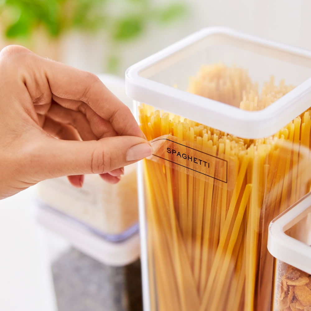 Iemand plakt een etiket uit de Rotho LOFT-stickerset – keukenorganisatie – met het opschrift SPAGHETTI op een doorzichtige bak met ongekookte spaghetti, terwijl op de achtergrond nog meer gelabelde voedselbakjes uit dezelfde serie te zien zijn.