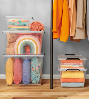 In de Orplast SimpleStore Box WHEEL (40 l, 58 x 39 x 25 cm) zitten kleurrijke kussens, een teddybeer, een regenboogkussen en dekens. Ernaast hangen gele en oranje kledingstukken. De stapelbare boxen staan netjes op de houten vloer.