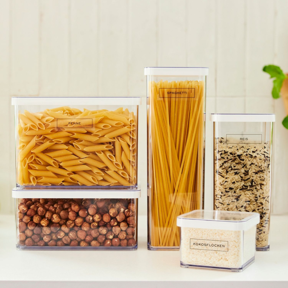Vijf doorzichtige plastic bakjes met penne, spaghetti, rijst, hazelnoten en kokosvlokken staan keurig op een lichte keukenbar, elk voorzien van een Rotho's LOFT-sticker – keukenorganisatie voor gemakkelijke herkenning.