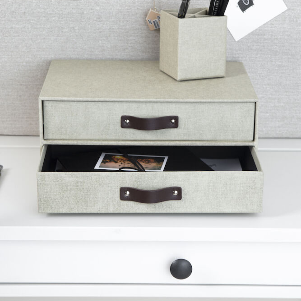 Op een wit bureau staat de Bigso BIRGER ladekast met 2 vakken van linnen canvas in lichtgrijs; de onderste lade is open en bevat papieren en een foto; ernaast staat een bijpassende pennenhouder tegen een lichte muur.