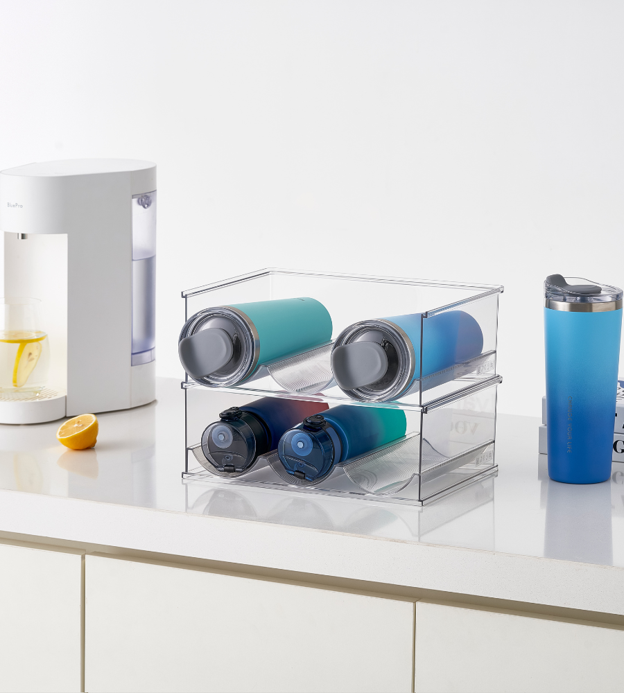 De BNB x VCN-fleshouder KLAR - set van 2 stuks houdt vier herbruikbare flessen netjes op een wit aanrecht, terwijl er een blauwe reisbeker, een doorgesneden citroen en een witte drinkbeker met citroenwater naast staan.