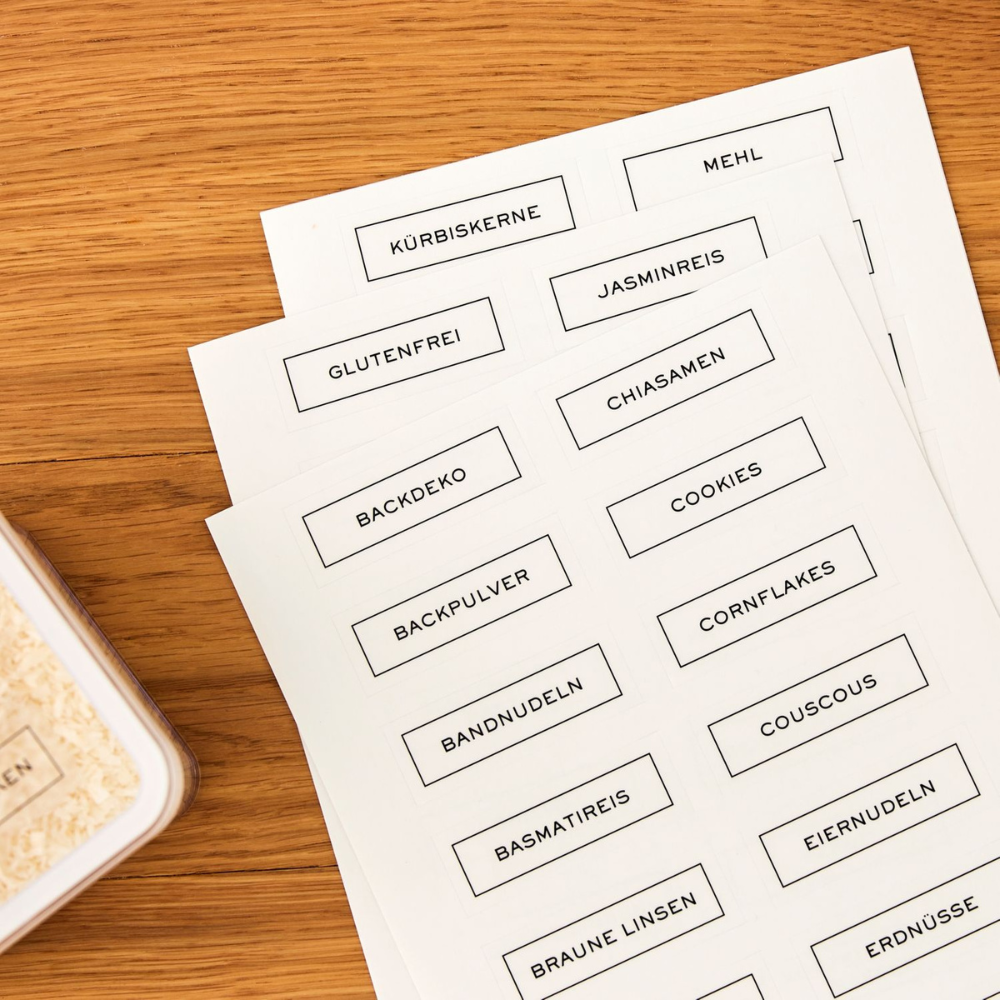 Drie vellen LOFT-stickers – keukenorganisatie van Rotho met Duitse namen van voedingsmiddelen zoals pompoenpitten, meel en jasmijnrijst, op hout naast voorraadbakjes – ideaal als etiketten voor droge voorraden.