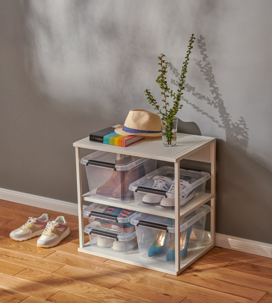 Op een witte plank staan Orplast SimpleStore Box - 39x29x11 cm (8,5 l) opbergdozen met schoenen erin. Bovenop staan een potplant, boeken en een strohoed. Twee paar sportschoenen staan op de houten vloer voor een grijze muur.