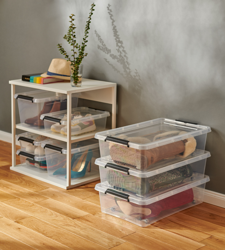 Drie BPA-vrije Orplast SimpleStore-boxen (39x29x16,5 cm, 12,5 l) staan vol met schoenen en zijn opgestapeld op een houten vloer, naast een klein wit rek met nog meer doorzichtige boxen, boeken, een hoed en een vaas met een groene plant.