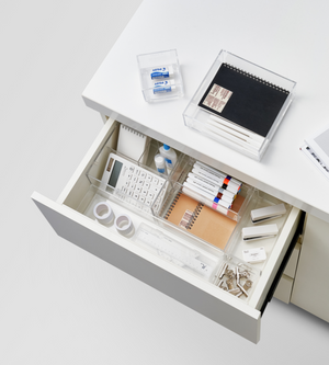 Een open witte bureaulade is netjes geordend met de SLIDE-lade-organizer KLAR - HOCH (BNB x VCN, verschillende maten) en biedt ruimte voor de belangrijkste kantoorbenodigdheden zoals rekenmachines, pennen, markeerstiften, plakband, paperclips, gummen en kleine notitieboekjes; andere spullen liggen op het bureau erboven.