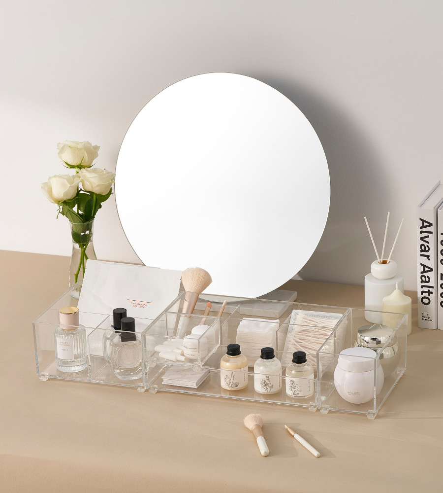 Een wastafel met een ronde spiegel, witte rozen in een vaas, huidverzorgingsproducten en make-up in de BNB x VCN SLIDE-lade-organizer KLAR - HOCH (verschillende maten), kwasten, wattenschijfjes, diffuser-staafjes en boeken aan de rechterkant – allemaal op een beige ondergrond.