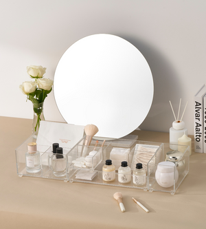 Een wastafel met een ronde spiegel, witte rozen in een vaas, huidverzorgingsproducten en make-up in de BNB x VCN SLIDE-lade-organizer KLAR - HOCH (verschillende maten), kwasten, wattenschijfjes, diffuser-staafjes en boeken aan de rechterkant – allemaal op een beige ondergrond.
