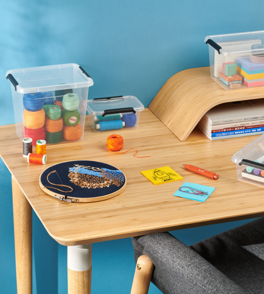 Een houten bureau met kleurrijk borduurgaren, de Orplast SimpleStore Box STACK&NEST (39x39x26 cm, 25 l), boeken, een borduurraam waar nog aan gewerkt wordt, schetsmatige post-its en een pen op een blauwe achtergrond. Een grijze stoel is gedeeltelijk zichtbaar.