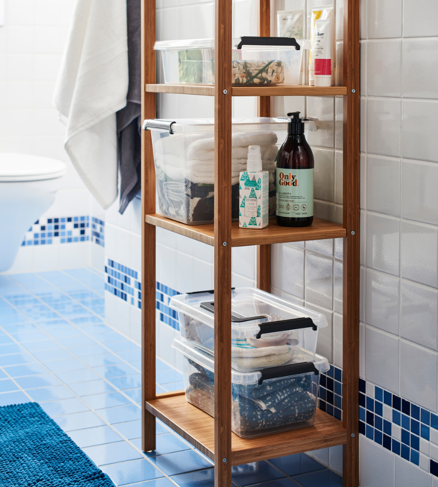 Een houten badkamerrek staat op blauwe tegels en bevat de Orplast SimpleStore-box – 39x29x21,5 cm (15,5 l), opgevouwen handdoeken, flessen en toiletartikelen. Een witte handdoek hangt naast een wit toilet en een betegelde muur op de achtergrond.