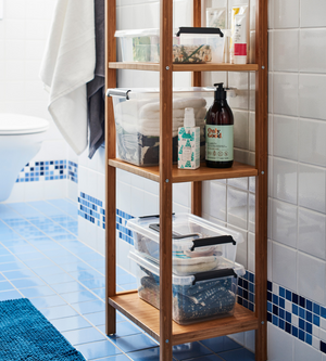 Een houten badkamerrek staat op blauwe tegels en bevat de Orplast SimpleStore-box – 39x29x21,5 cm (15,5 l), opgevouwen handdoeken, flessen en toiletartikelen. Een witte handdoek hangt naast een wit toilet en een betegelde muur op de achtergrond.