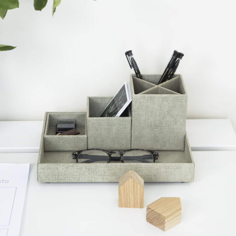 De LENA-bureauorganizer – linnen canvas van Bigso in grijs biedt ruimte voor pennen, een telefoon en nog veel meer. Houten blokjes en papier liggen op het witte bureau, terwijl er vanuit de hoek een groene plant tevoorschijn komt – ideaal om je bureau opgeruimd te houden.