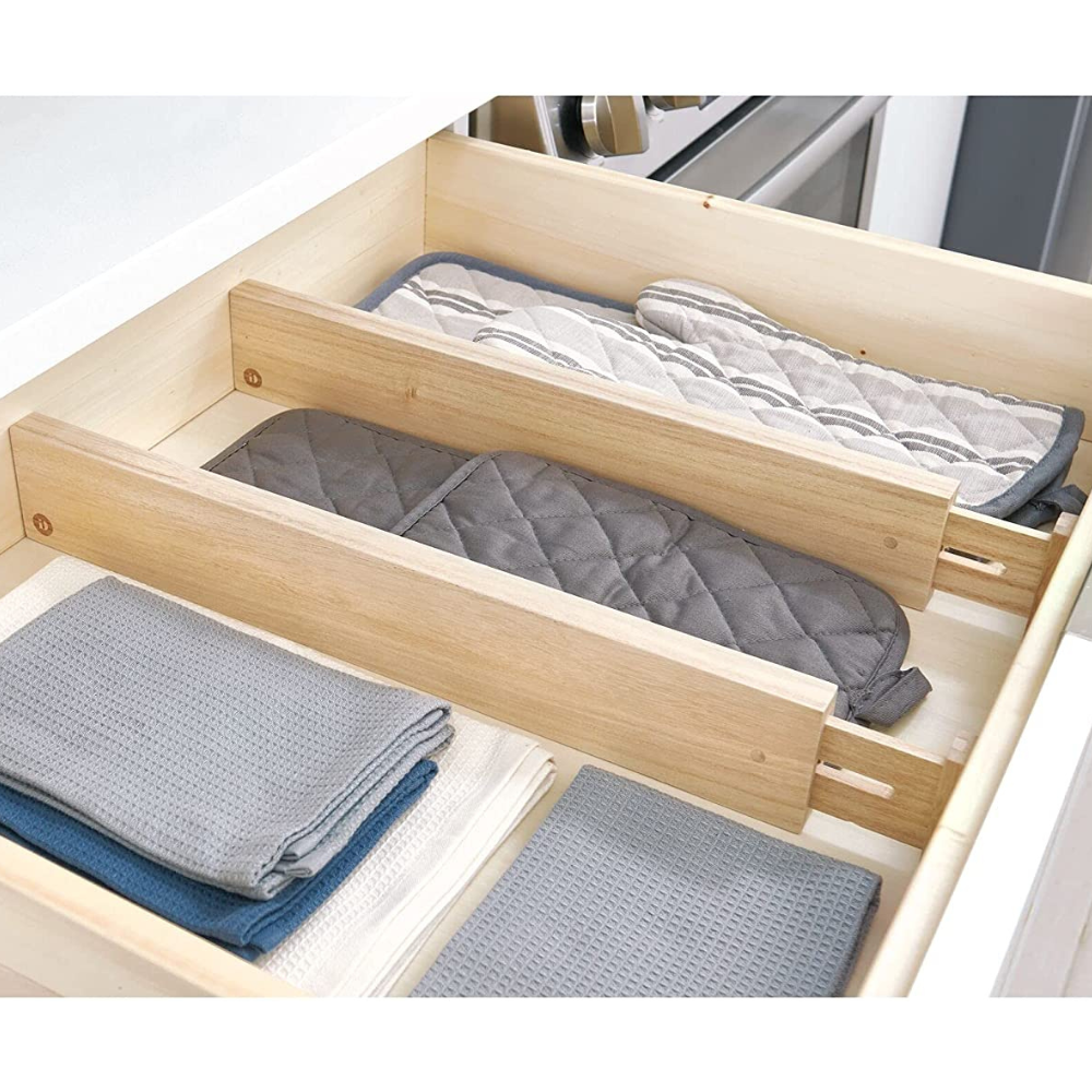 Een keukenlade met iDesign ECO - lage ladeverdelers WOOD (set van 2 stuks), waarmee grijze en beige pannenlappen en opgevouwen theedoeken overzichtelijk in aparte vakken worden opgeborgen.