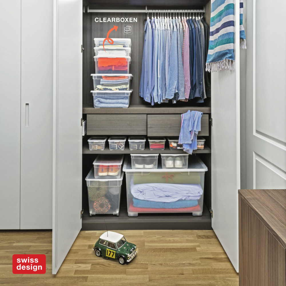 Een overzichtelijke kledingkast met de Rotho opbergbox CLEAR 46L (55x37,5x32 cm), hangende overhemden, opgevouwen kleding en lades. Op de houten bodem staat een speelgoedauto. De kast is voorzien van een CLEARBOXEN-label en een Zwitsers designlogo in de hoek.