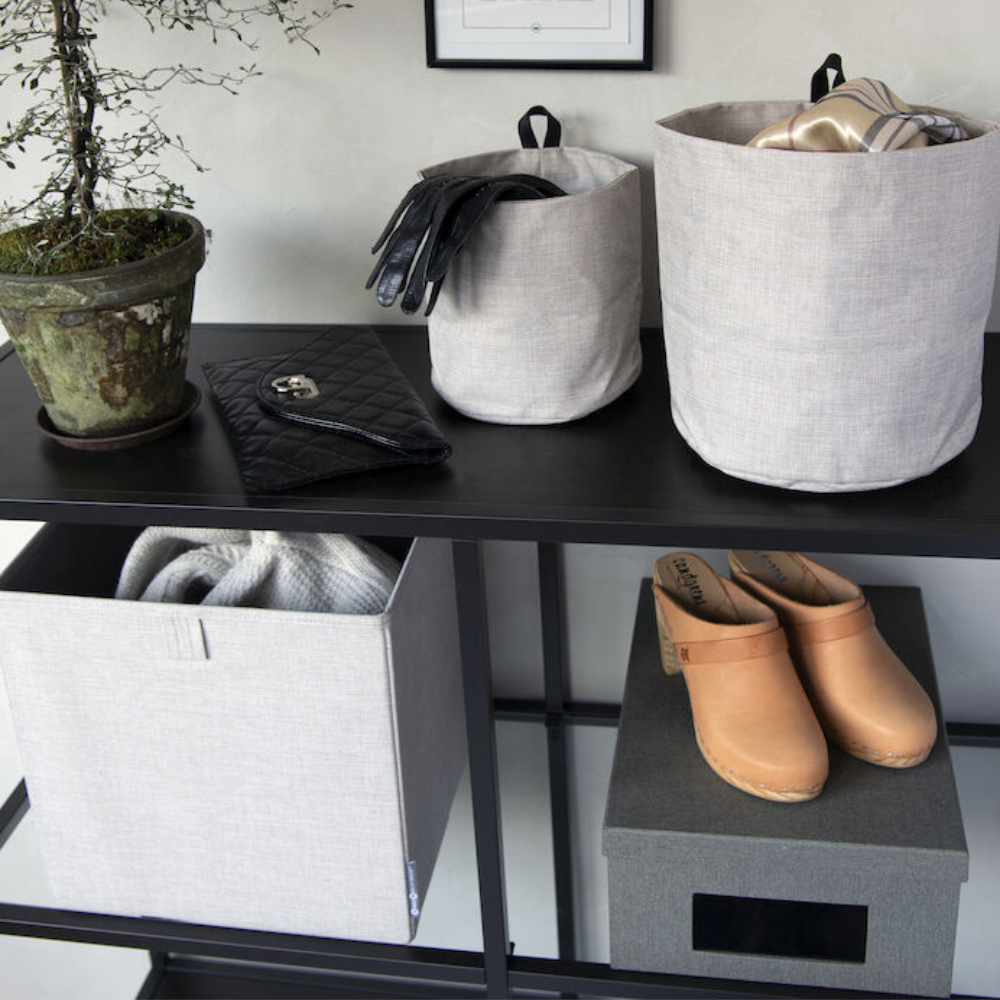 Op een zwarte plank staan een potplant, een zwarte handtas, twee Bigso SOFT opbergdozen CUBE – beige voor handschoenen en sjaals, lichtbruine klompen en een donkere opbergdoos. De neutrale tinten van de beige dozen zorgen voor een opgeruimde, georganiseerde look.