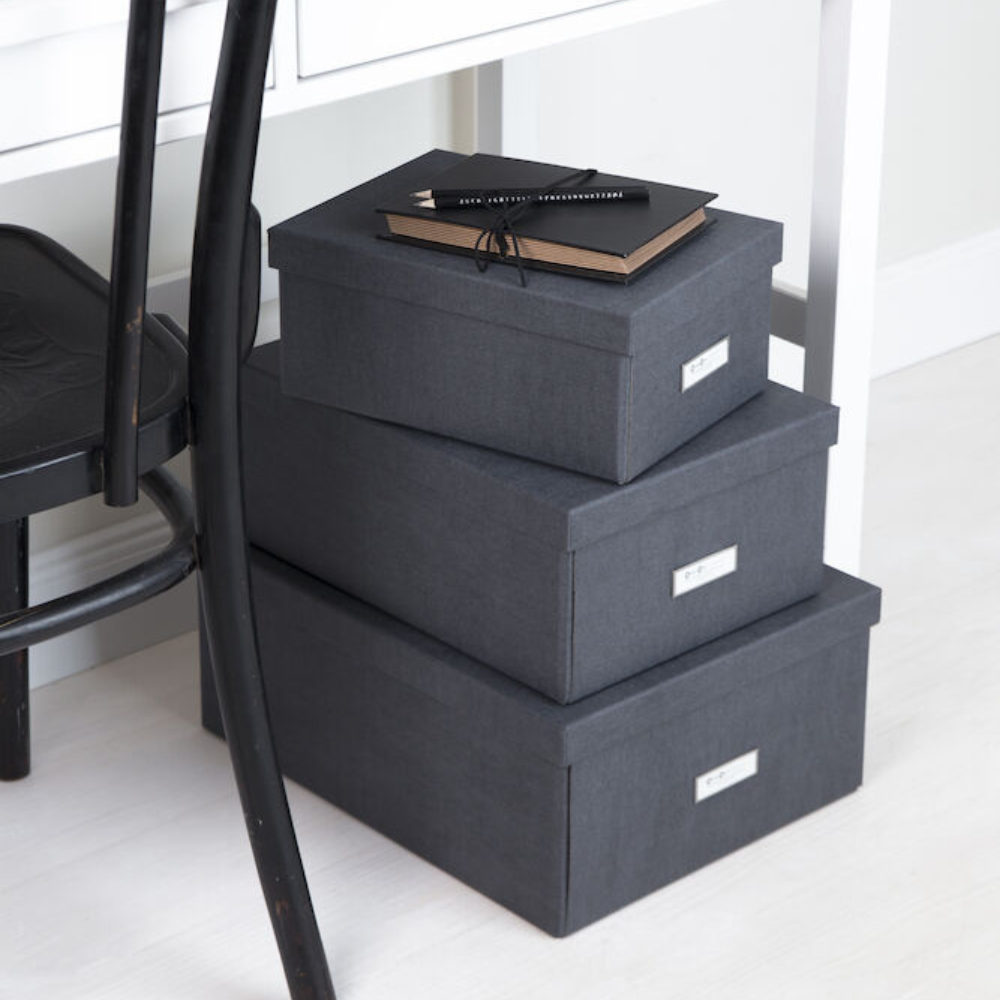 Drie stapelbare Bigso KARIN-opbergboxen van zwart zeildoek met etikethouders staan op een lichte vloer naast een zwarte stoel en een wit bureau. Op de bovenste box liggen twee notitieboekjes en een potlood.