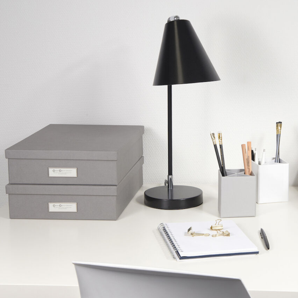 Een opgeruimd bureau met twee gestapelde Bigso OSKAR opbergboxen A4 in grijs, een zwarte bureaulamp, een notitieboekje met gouden paperclips en bakjes voor potloden en balpennen, allemaal tegen een witte muur.