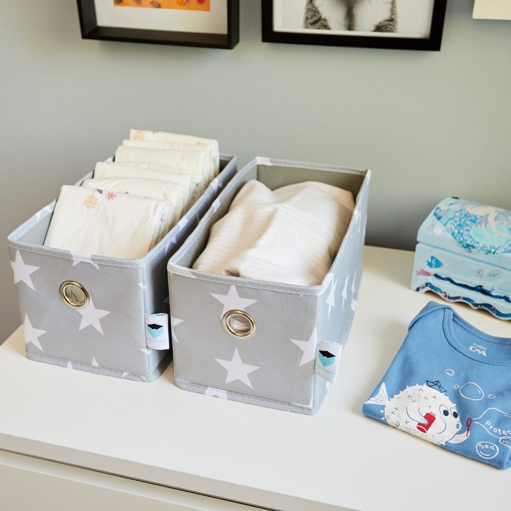 Two STAR Organizer smal grijs - set van 2 stuks van Love it Store it, met witte sterren, om babyluiers en kleding op een commode op te bergen. Op de commode staan ook een blauwe tissuebox en een opgevouwen blauw rompertje, waardoor er een handige organizer ontstaat onder de wanddecoratie.