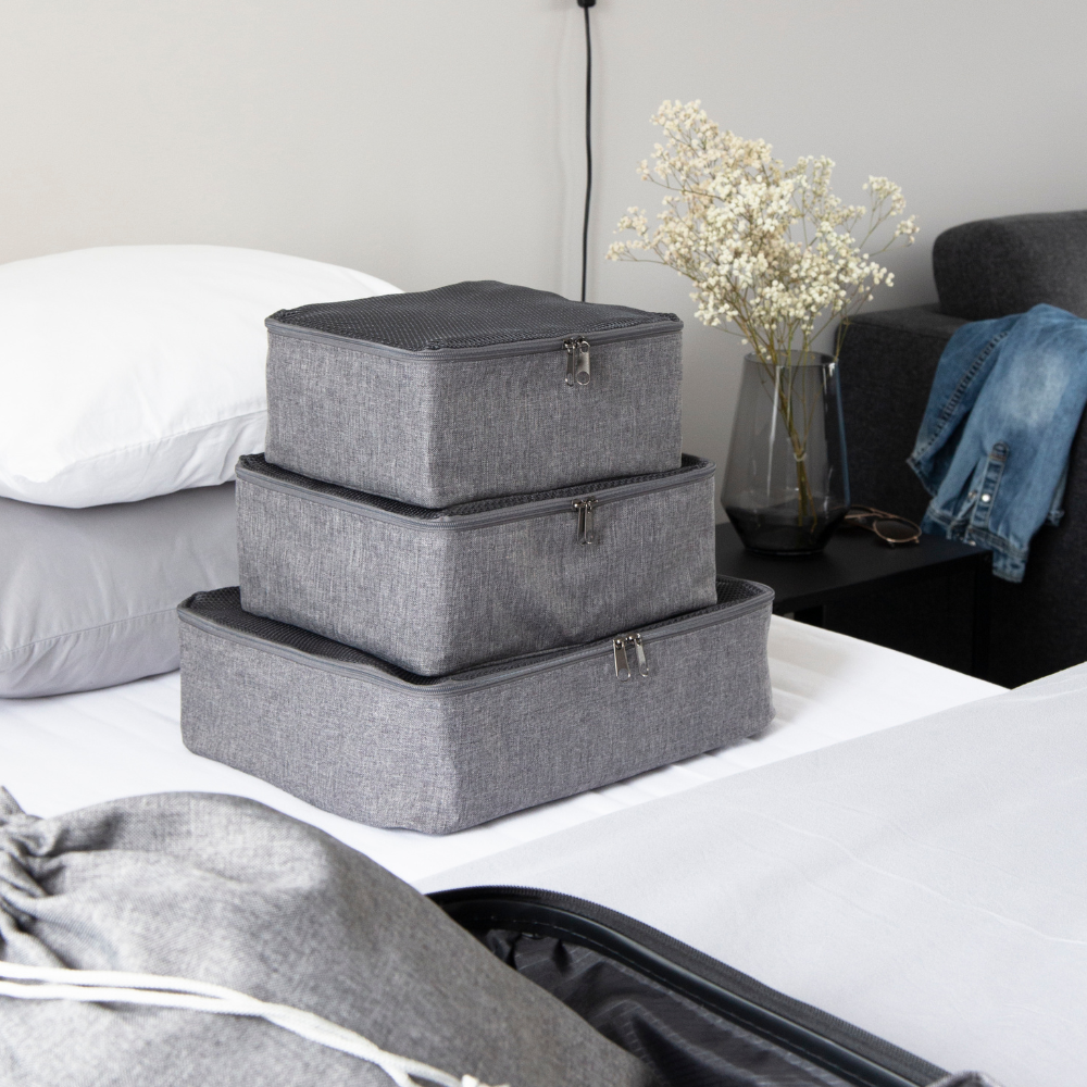 Drie Bigso TRAVEL Organizer S-kubussen in grijs liggen opgestapeld op een net opgemaakt bed. Op het nachtkastje liggen gedroogde bloemen, een bril, een spijkerbroek en stukjes van een kussen en een koffer – ideaal voor iedereen die zijn koffer wil organiseren.