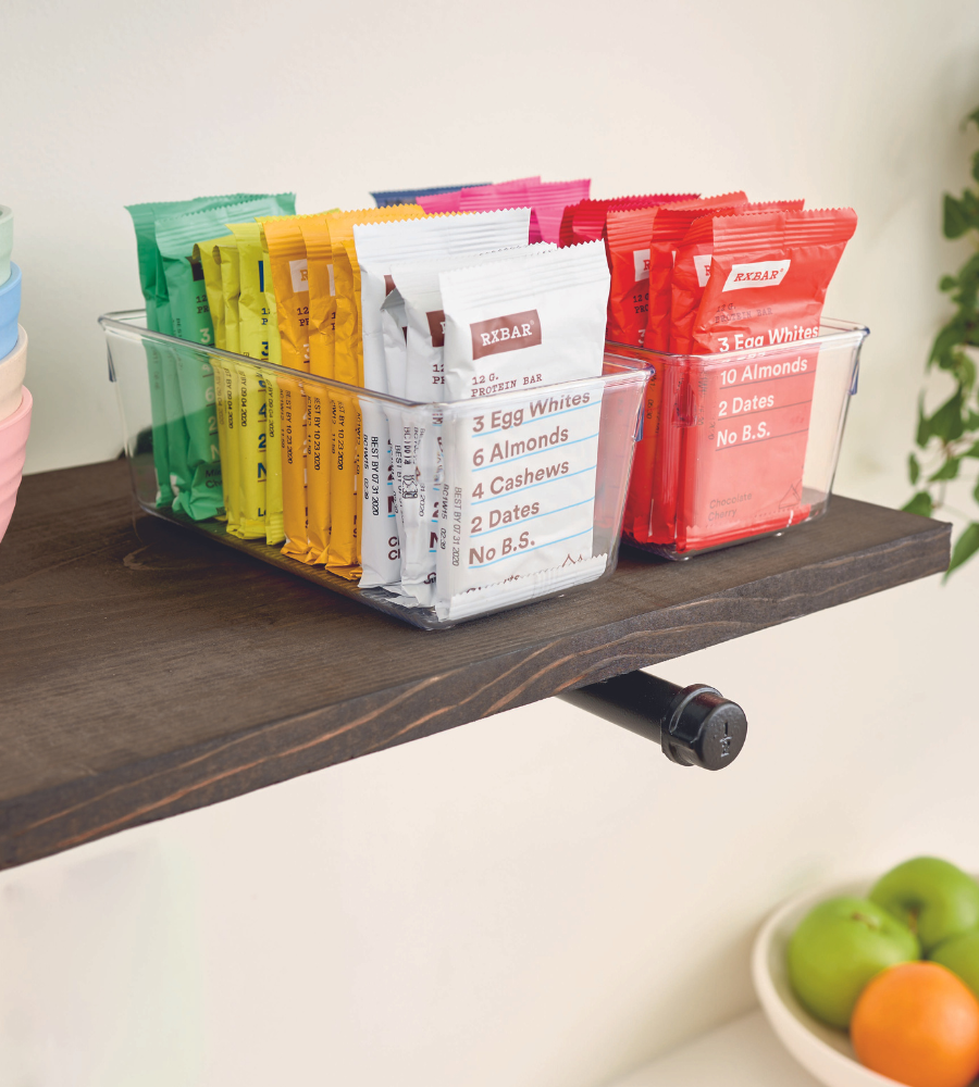 Een iDesign LINUS - lade-indeler HOOG houdt kleurrijke RXBAR-eiwitrepen op een donkere houten plank; daaronder zie je een schaal met fruit.