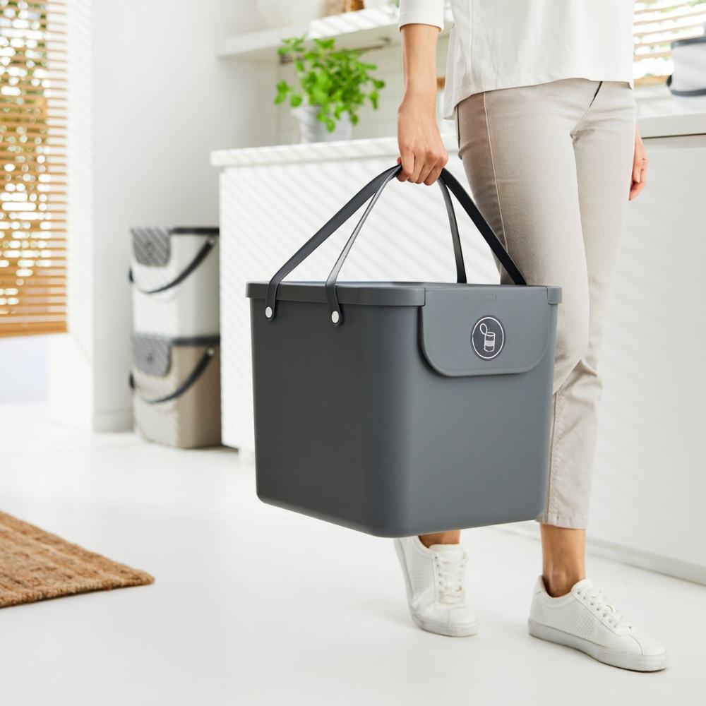 Iemand in lichte kleding en witte sneakers draagt een groot Rotho ALBULA-afvalsysteem van 40 liter – antraciet met handvatten – in een lichte, moderne keuken. Op de achtergrond zie je planten en keukengerei.