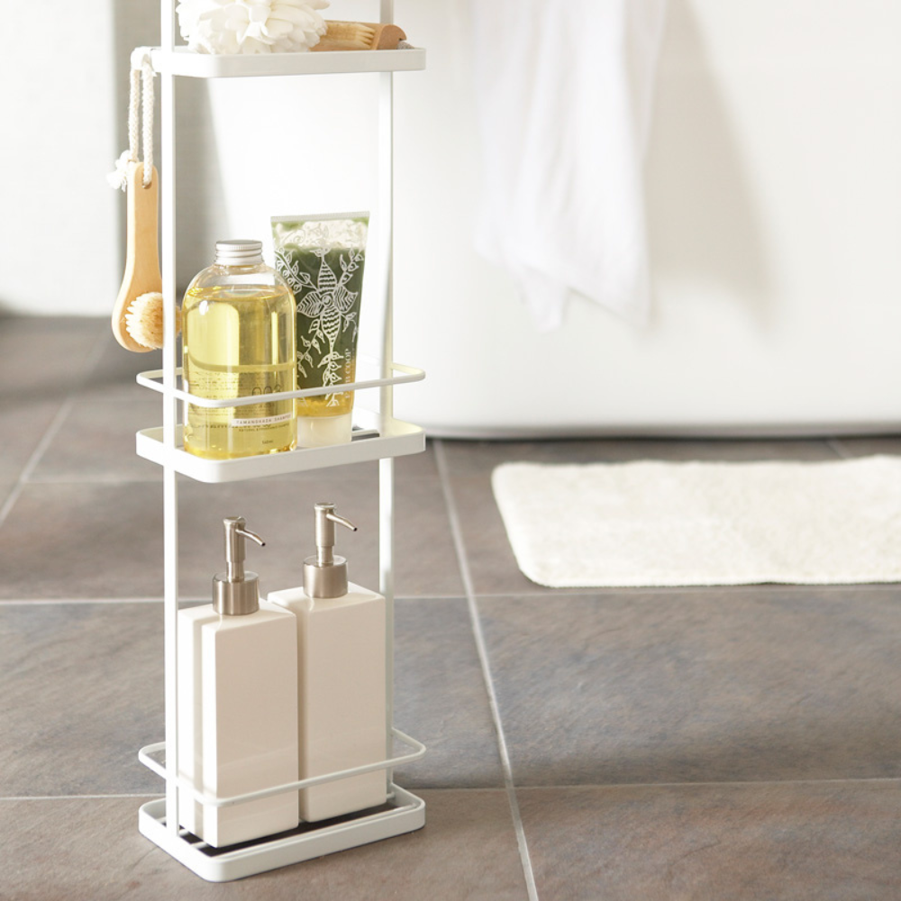 De witte YAMAZAKI TOWER badkamerplank biedt extra opbergruimte en zorgt ervoor dat zeep, lotion, potjes, borstels en sponsjes netjes op een grijze tegelvloer naast het witte bad en het badmatje staan.