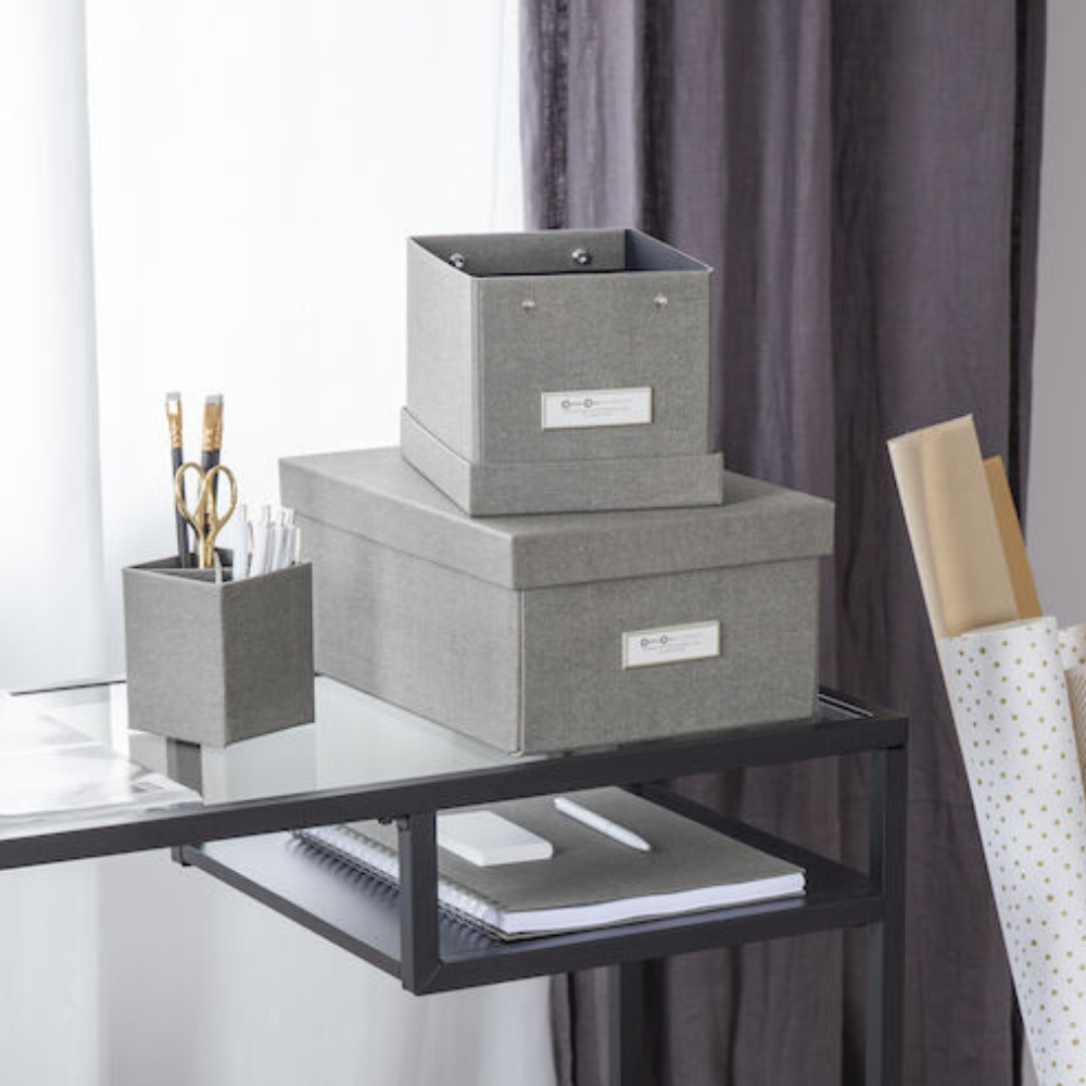 Een modern, opgeruimd bureau met de Bigso KARIN opbergdoos van grijs zeildoek met een etikettenhouder, een pennenhouder met een schaar en potloden, een notitieboekje en opgerold inpakpapier voor een minimalistische look.