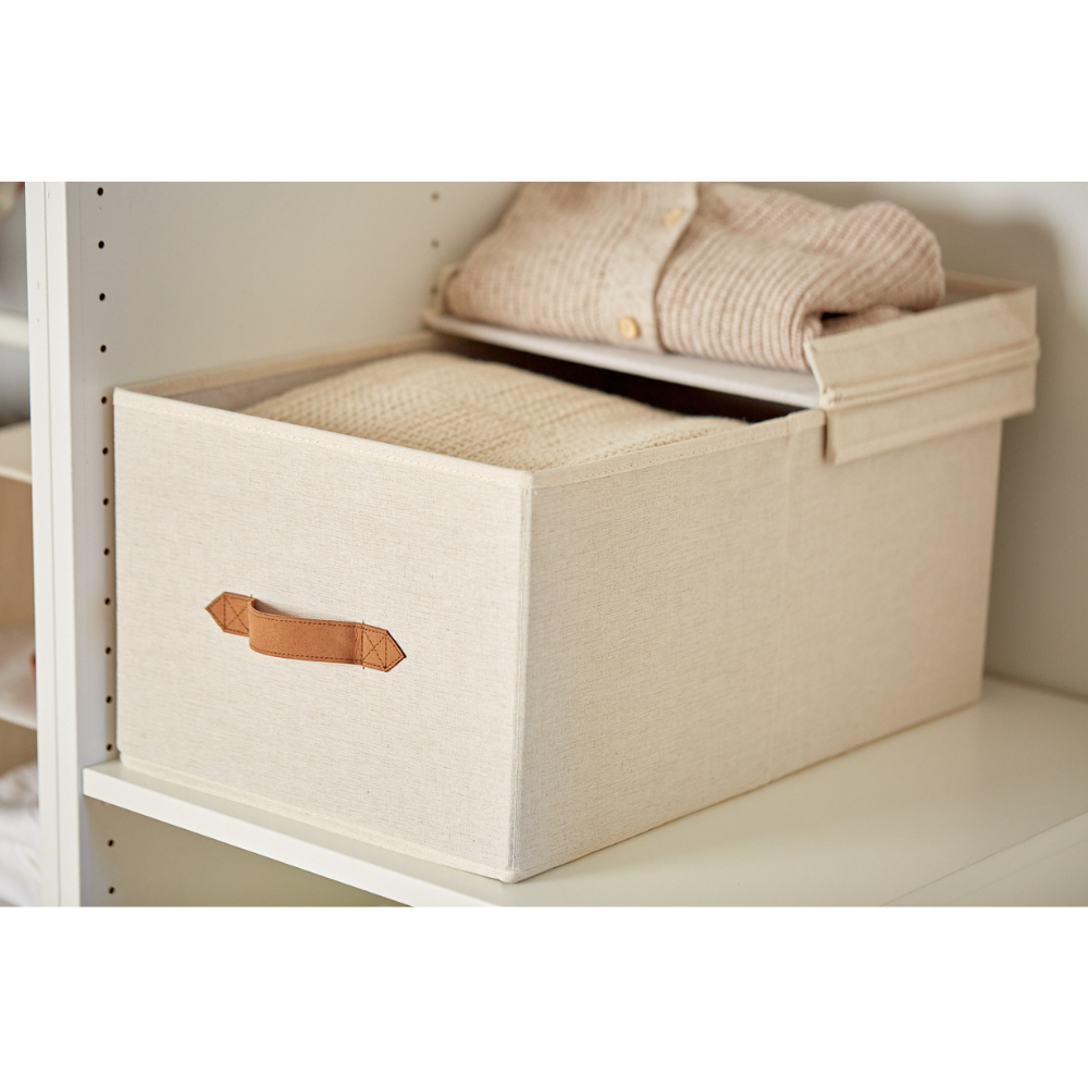 De PREMIUM opbergdoos met klapdeksel – beige van Love it Store it met bruine handgreep staat op een witte plank, waarvan het deksel gedeeltelijk openstaat en waarin netjes opgevouwen crèmekleurige truien liggen.