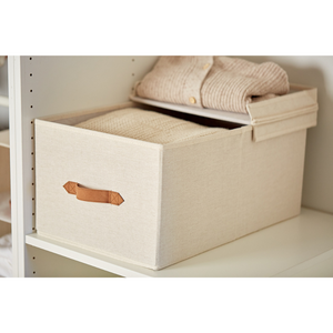De PREMIUM opbergdoos met klapdeksel – beige van Love it Store it met bruine handgreep staat op een witte plank, waarvan het deksel gedeeltelijk openstaat en waarin netjes opgevouwen crèmekleurige truien liggen.