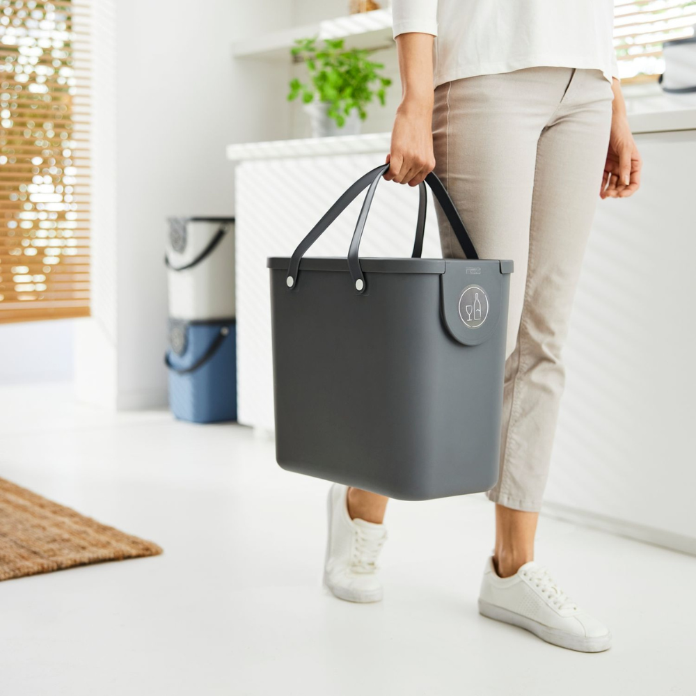 Iemand in een lichte broek en witte sneakers draagt een grote antracietkleurige ALBULA Recycling-afvalbak van 25 liter van Rotho met handvatten in een lichte, moderne ruimte vol planten en natuurlijk licht.