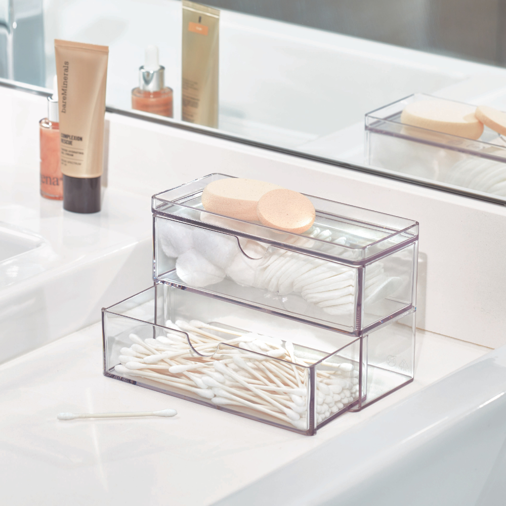 Twee stapelbare iDesign HOME EDIT - cosmetica-organizers in transparant met een lade staan op de badkamerwastafel en bieden ruimte aan wattenschijfjes, wattenstaafjes en sponsjes. Een tube huidverzorgingsproduct en een flesje staan ernaast bij de spiegel.