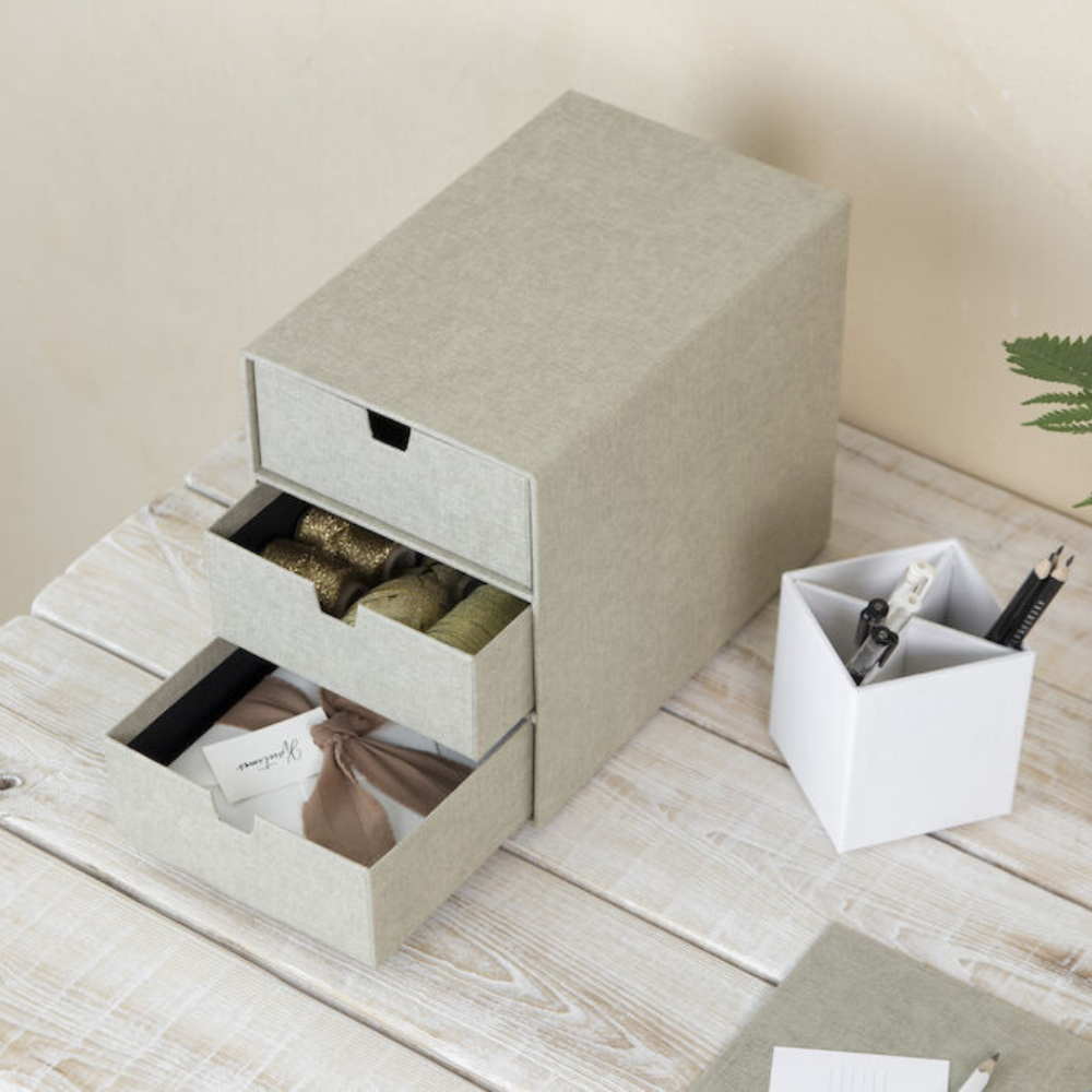 De grijze Bigso INGRID-ladekast met 3 vakken – linnen canvas – staat op een witte houten tafel; in de open lades liggen linten, stoffen en cadeaulabels – perfect voor het organiseren van je kantoor. Ernaast staat een witte pennenhouder voor pennen en scharen.