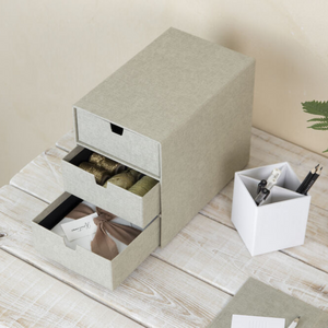 De grijze Bigso INGRID-ladekast met 3 vakken – linnen canvas – staat op een witte houten tafel; in de open lades liggen linten, stoffen en cadeaulabels – perfect voor het organiseren van je kantoor. Ernaast staat een witte pennenhouder voor pennen en scharen.