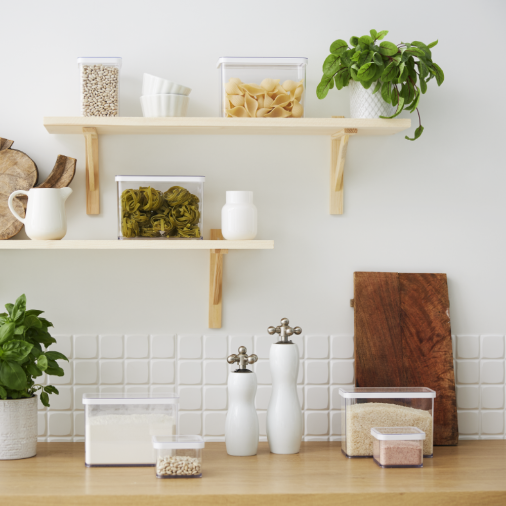 Op een modern keukenblad staat de Rotho-voorraadbussenset LOFT (6-delig) met pasta, granen en bonen overzichtelijk opgesteld. Daarboven bevinden zich houten planken met nog meer voorraadbussen en servies. Witte tegels en lichte houten accenten maken de frisse look helemaal af.