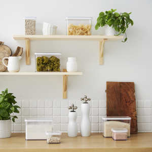Op een modern keukenblad staat de Rotho-voorraadbussenset LOFT (6-delig) met pasta, granen en bonen overzichtelijk opgesteld. Daarboven bevinden zich houten planken met nog meer voorraadbussen en servies. Witte tegels en lichte houten accenten maken de frisse look helemaal af.