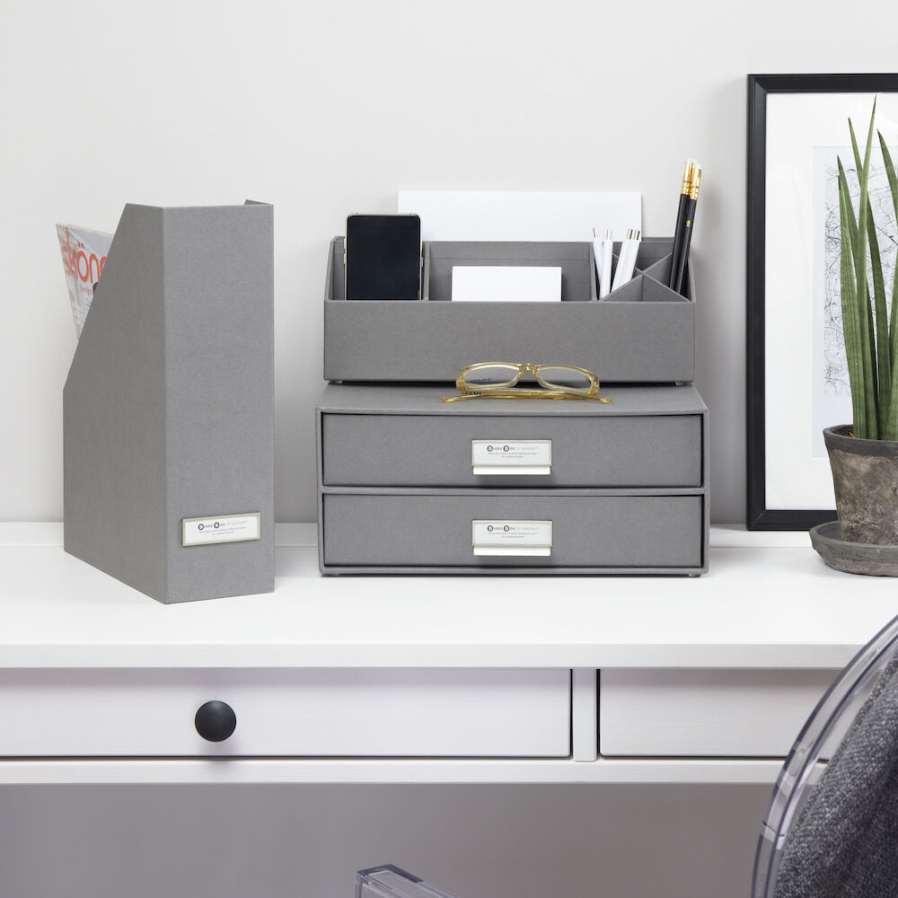 Een strak wit bureau met grijze kantooraccessoires, waaronder de Bigso VIKTORIA staande documenthouder – grijs met etikettenhouder, een A4-documentenhouder, twee gestapelde laden, glazen, een plant en ingelijste kunst tegen een witte muur.