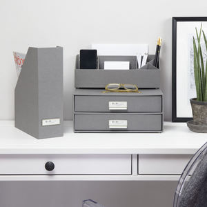 Een strak wit bureau met grijze kantooraccessoires, waaronder de Bigso VIKTORIA staande documenthouder – grijs met etikettenhouder, een A4-documentenhouder, twee gestapelde laden, glazen, een plant en ingelijste kunst tegen een witte muur.