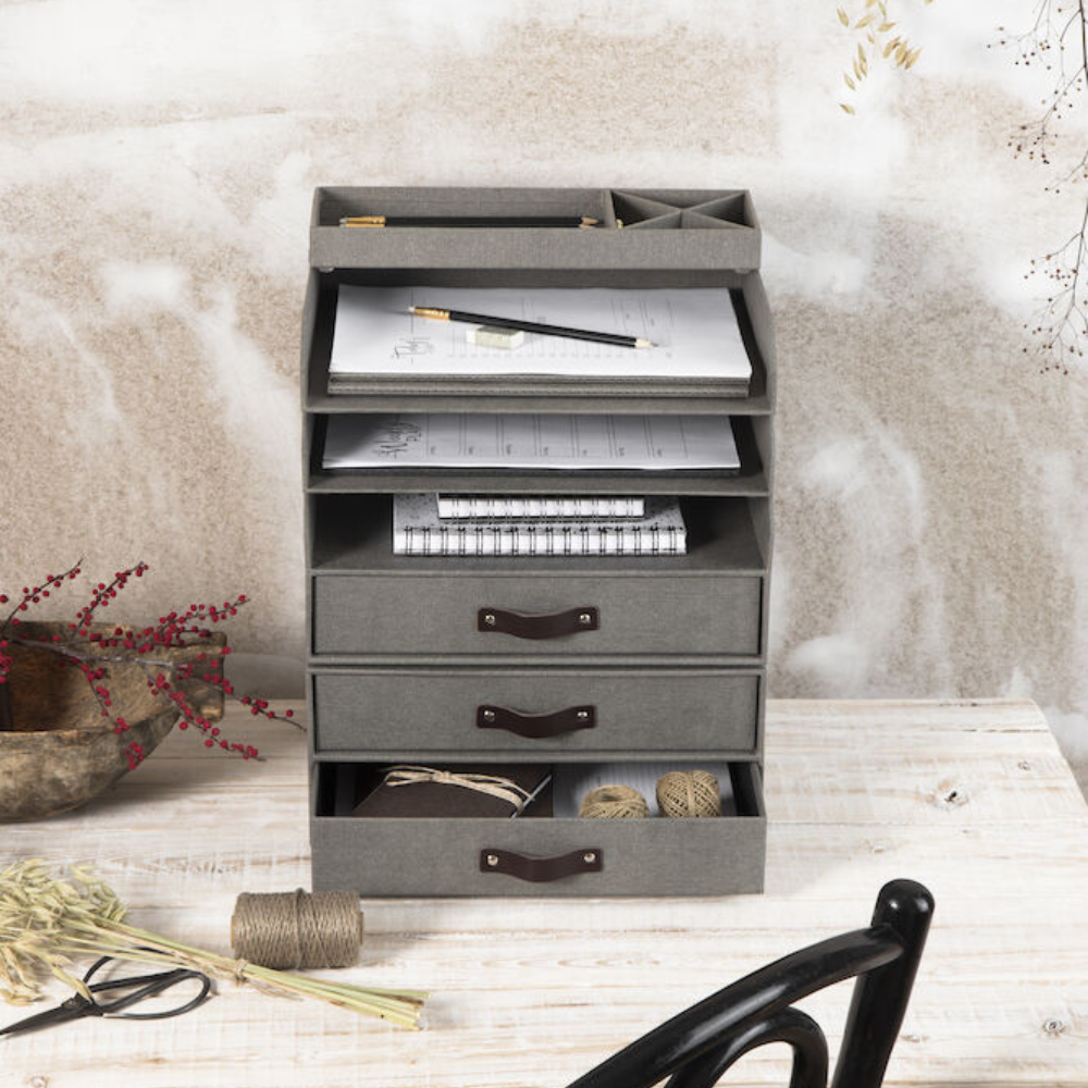 De Bigso BIRGER ladekast met 2 vakken in grijs canvas, met vakken en lades voor schriften, papieren, pennen en touwtjes, staat op een witte houten tafel naast gedroogde bloemen, een klos garen en een zwarte stoel.