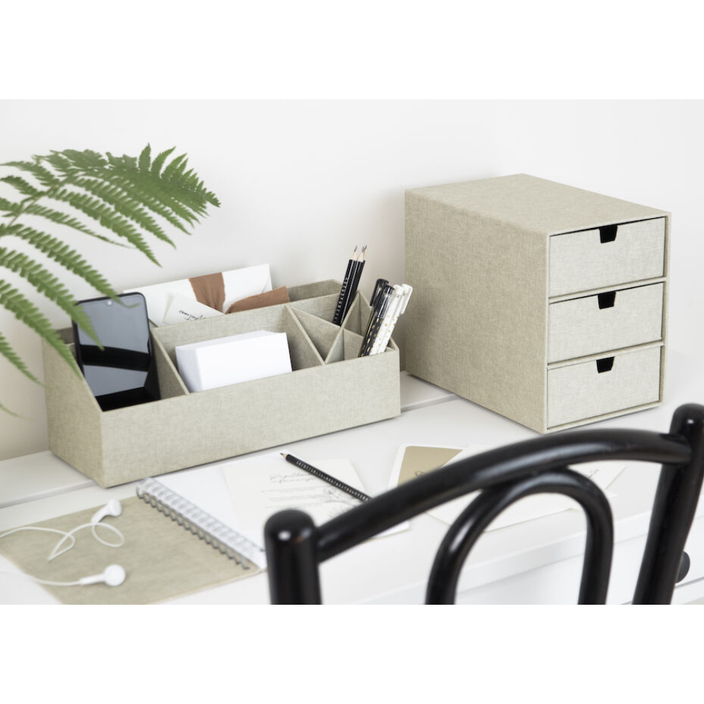 Een opgeruimd bureau met de ELISA-bureauorganizer – linnen canvas van Bigso in beige, waarin je je telefoon, enveloppen, pennen en een schaar kwijt kunt, naast een bijpassend kastje met drie lades. Varenbladeren, een notitieboekje, een koptelefoon en een bril maken het opgeruimde witte bureau compleet.