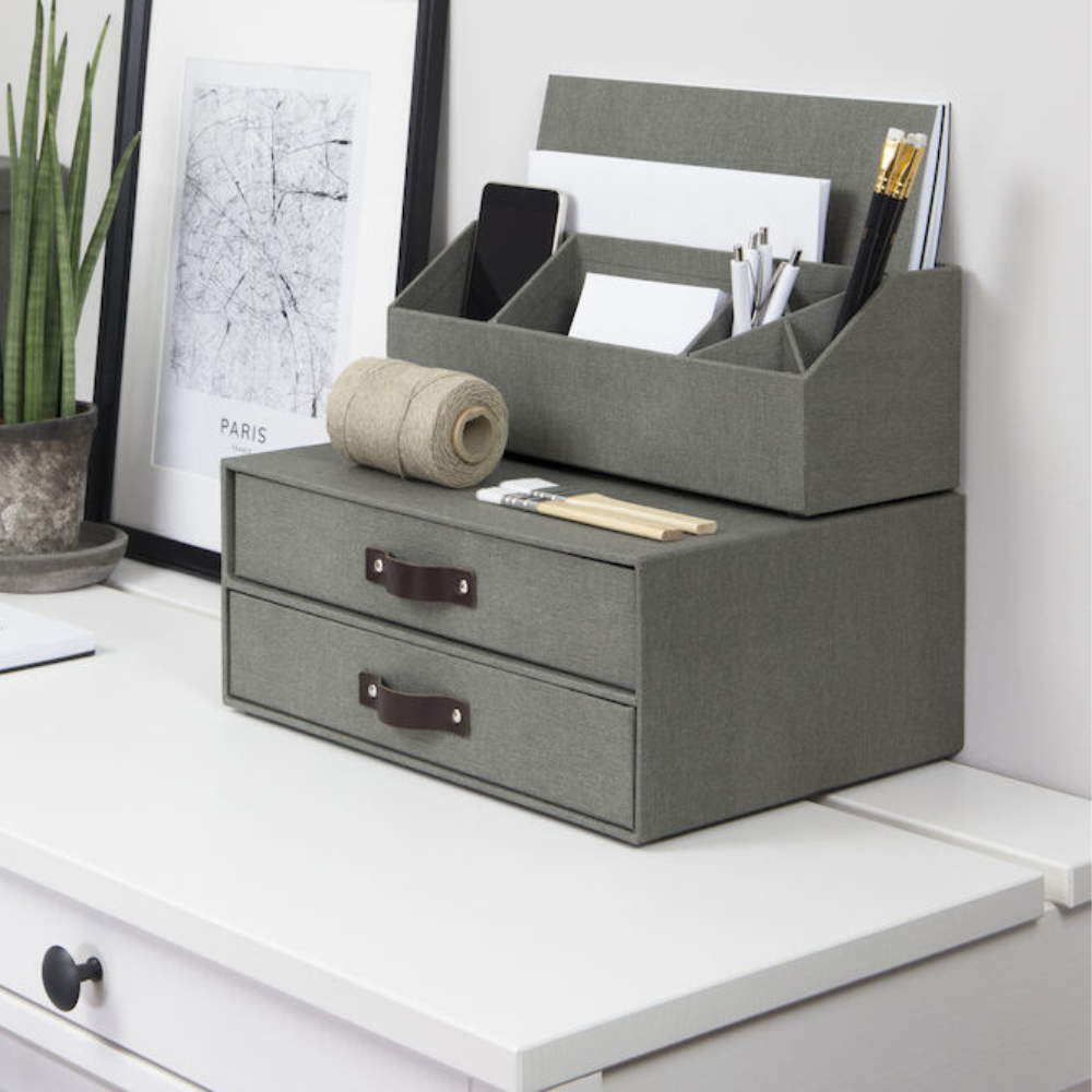 Op een opgeruimd bureau staat de Bigso BIRGER-ladekast met 2 vakken van grijs canvas, waarin enveloppen, pennen, scharen en een telefoon passen. Ernaast staan een potplant, een ingelijste afbeelding van Parijs en een rol bindgaren op het witte oppervlak.