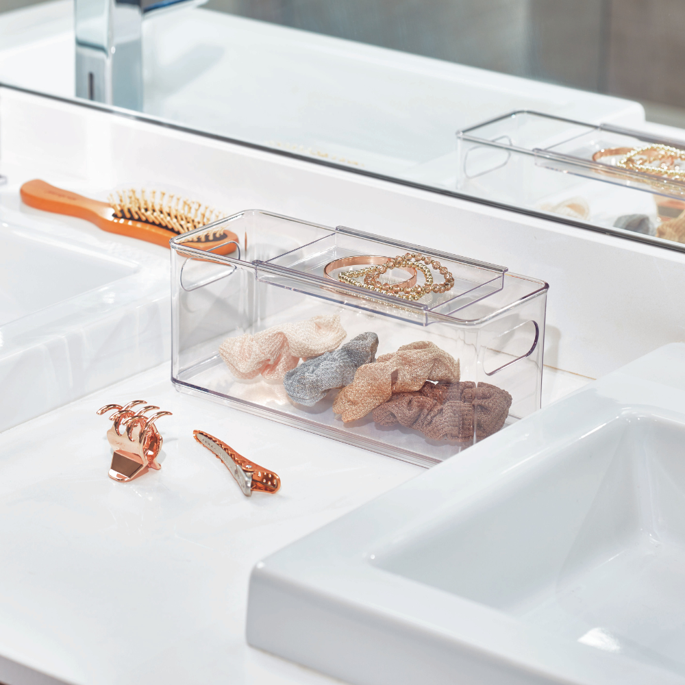 Een iDesign HOME EDIT-opbergbakje – Accessory staat op een wit badkamertafeltje, waarin haarelastiekjes en sieraden netjes worden opgeborgen. Vlakbij liggen een gouden haarspeld, een kam, twee wasbakken en een spiegel die de opgeruimde ruimte weerspiegelt.