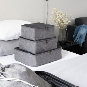 Drie grijze Bigso TRAVEL Organizer M’s liggen op een net opgemaakt bed, gestapeld naast een open koffer. Een nachtkastje met gedroogde bloemen, een bril en een opgevouwen overhemd maken de perfecte omgeving voor deze veelzijdige spullen compleet.
