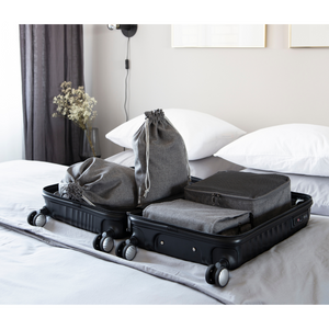 Een zwarte koffer staat open op een opgeruimd bed, gevuld met grijze opbergkubussen, zakjes en de Bigso TRAVEL waszak Grijs - 2 stuks - ideaal voor elke reis. Gedempt licht, witte kussens en een minimalistische inrichting maken het plaatje compleet.