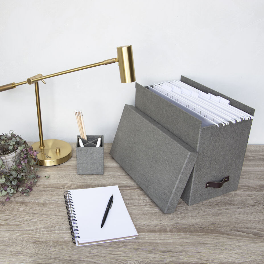Een modern bureau met een gouden lamp, een plantje, een pennenhouder, een openliggend notitieboekje en een Bigso JOHAN hangmappenbox van grijs zeildoek om je A4-papieren netjes te ordenen op een houten oppervlak.