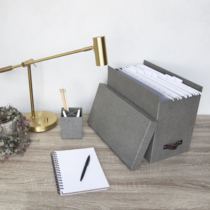 Een modern bureau met een gouden lamp, een plantje, een pennenhouder, een openliggend notitieboekje en een Bigso JOHAN hangmappenbox van grijs zeildoek om je A4-papieren netjes te ordenen op een houten oppervlak.