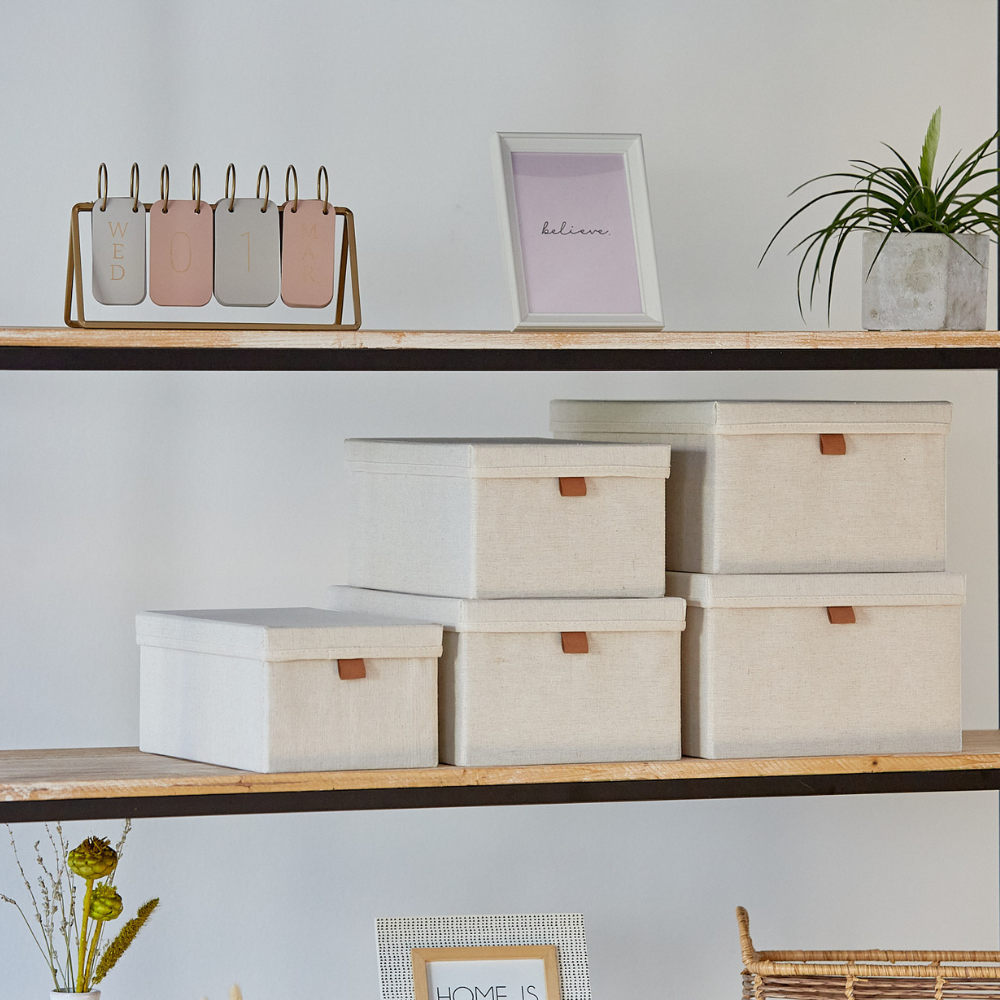 Een houten plank met Love it Store it PREMIUM opbergdozen met deksel in beige (set van 5), verstevigde kartonnen wanden, een ingelijste print met "believe", een metalen kalender op woensdag, een potplant en gedroogde bloemen – stijlvol georganiseerd in huis.