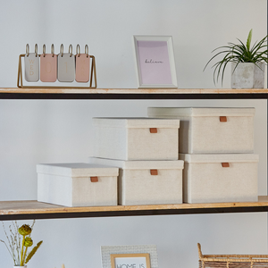 Een houten plank met Love it Store it PREMIUM opbergdozen met deksel in beige (set van 5), verstevigde kartonnen wanden, een ingelijste print met "believe", een metalen kalender op woensdag, een potplant en gedroogde bloemen – stijlvol georganiseerd in huis.