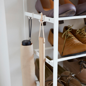 Een close-up van de YAMAZAKI TOWER schoenenrek van hout - wit, met bruine en lichtbruine schoenen erop. Aan de haken aan de zijkant hangen een beige paraplu en een houten schoenlepel, wat dit ruimtebesparende ontwerp zowel praktisch als stijlvol maakt.